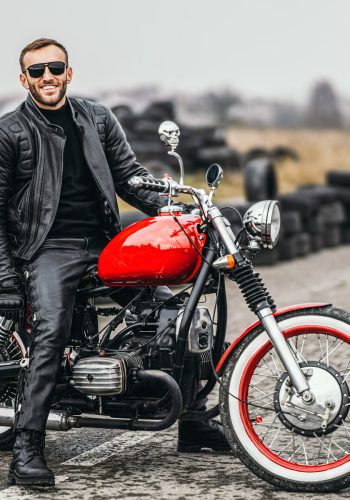Red motorbike with rider. A man in a black leather jacket and pa Red motorbike with rider. A man in a black leather jacket and pants stands sideways in the middle of the road. Tires are laid on the background.